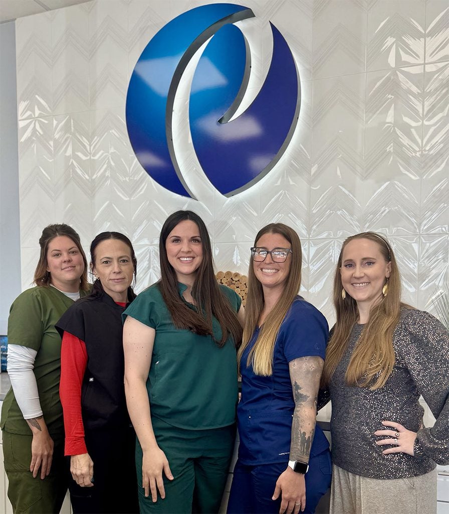 Primary-health-partners-broken-arrow - Dr. Ashley Pojezny, Do - Primary Care Physician Five women in scrubs stand together in a clinic lobby, posing in front of a large blue circular logo on a white patterned wall.